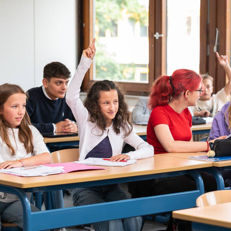 Mehrere Schülerinnen und Schüler sitzen in einem Klassenzimmer an Tischen, wobei einige die Hand heben, während andere sich unterhalten.