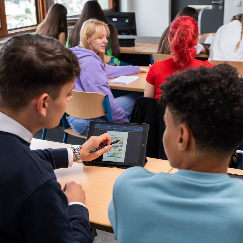 Schüler in einem Klassenzimmer arbeiten gemeinsam an Tablets, während einige nach vorne schauen.