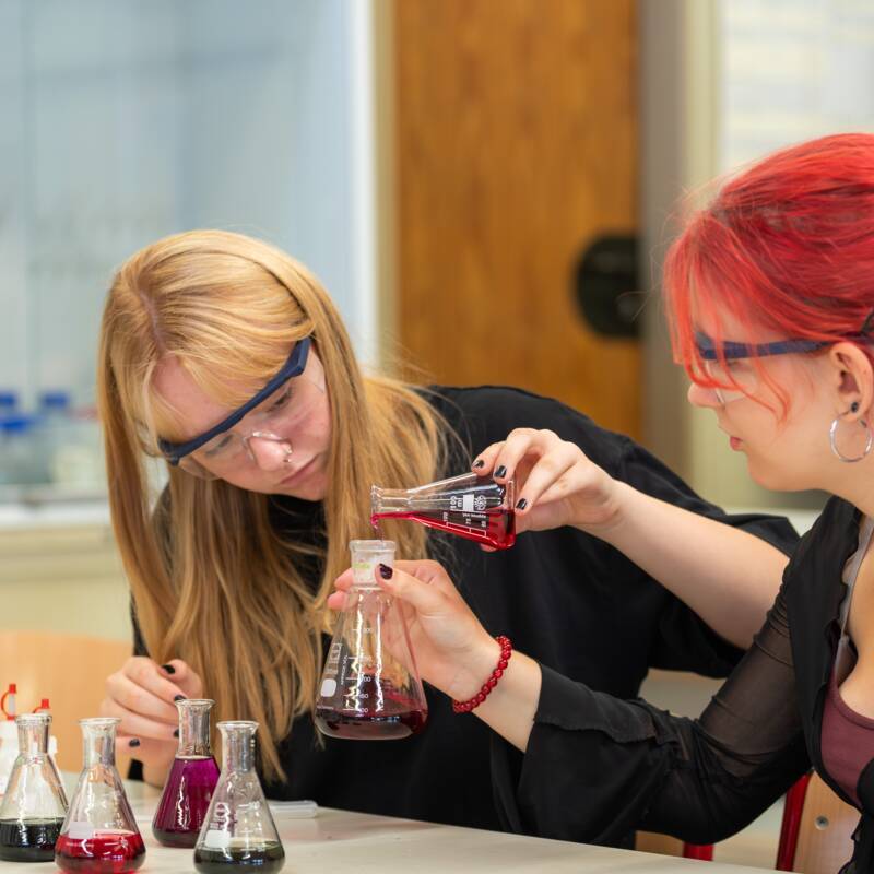 Zwei Schülerinnen mit Schutzbrillen befinden sich im Chemiesaal und arbeiten mit bunten Flüssigkeiten in Erlenmeyerkolben.