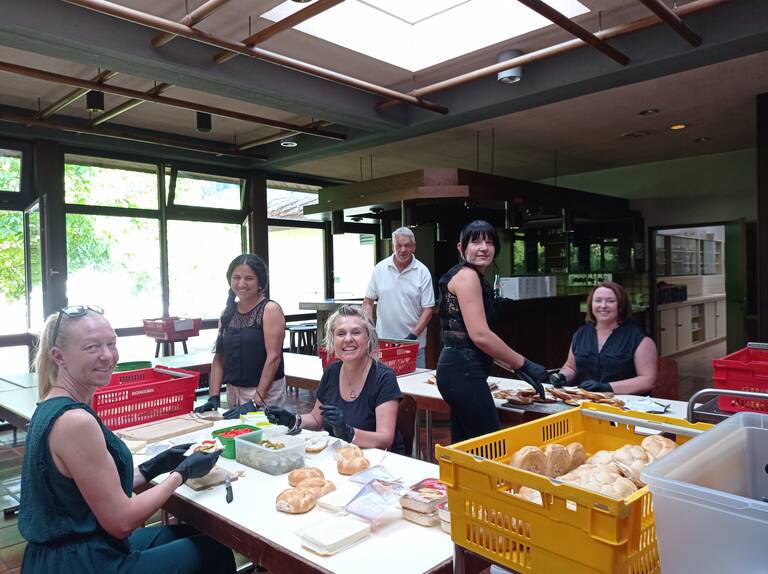 Eine Gruppe von sechs Personen bereitet an einem Tisch in einem hellen Raum Sandwiches vor, umgeben von Brot und Kisten.