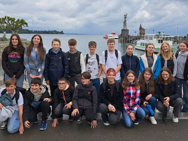 Eine Gruppe von jungen Menschen posiert für ein Foto vor einem See mit einer Statue und einem Boot im Hintergrund.
