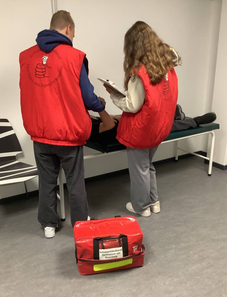 Zwei Schüler in roten Westen stehen vor einer Liege, auf dem jemand liegt, und tragen Klemmbretter, während eine rote Sani-Tasche auf dem Boden steht.