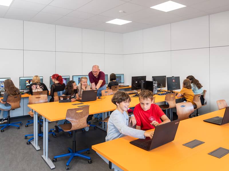 In einem hellen Klassenraum arbeiten Kinder und ein Erwachsener gemeinsam an Computern und Laptops.