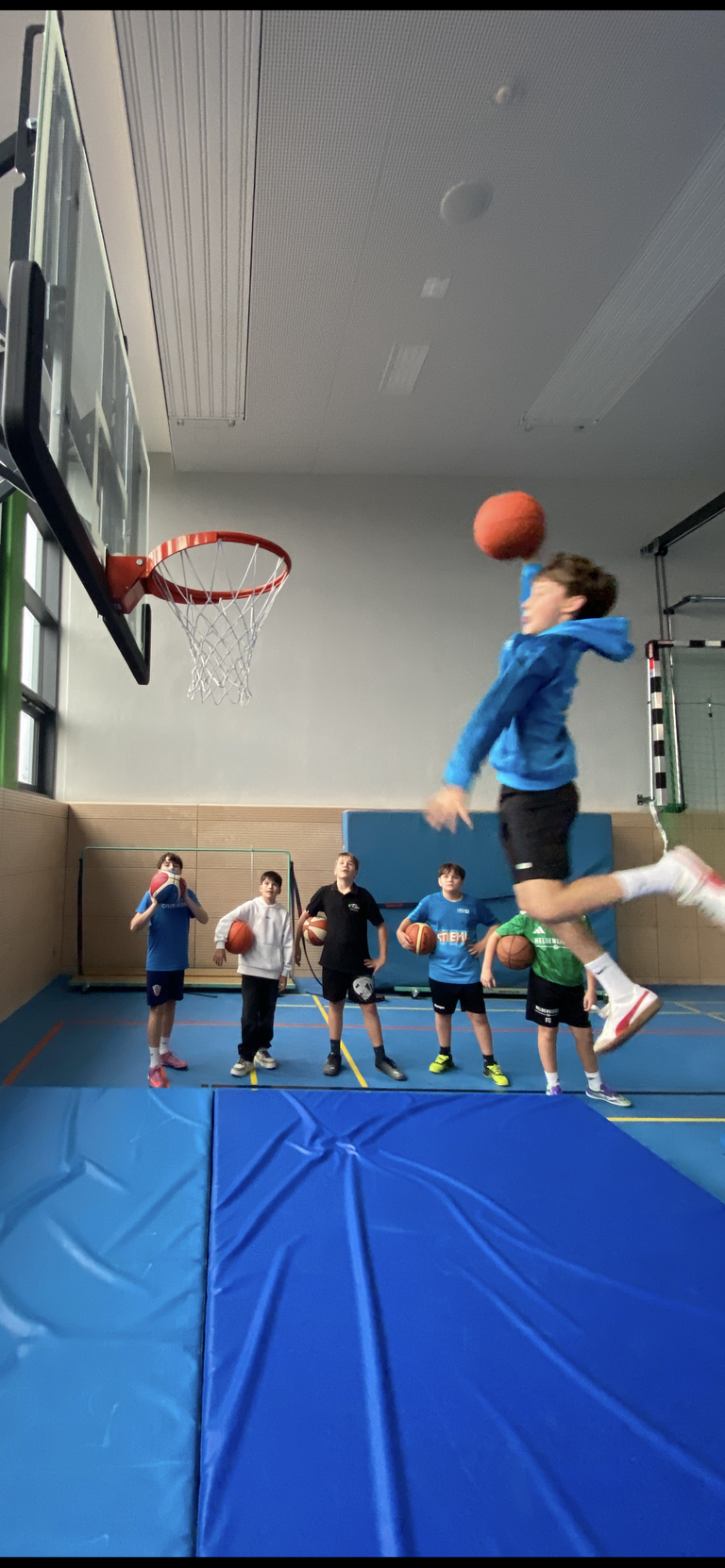 Schüler wirft einen Ball in den Basketballkorb, andere Schüler schauen im Hintergrund zu.