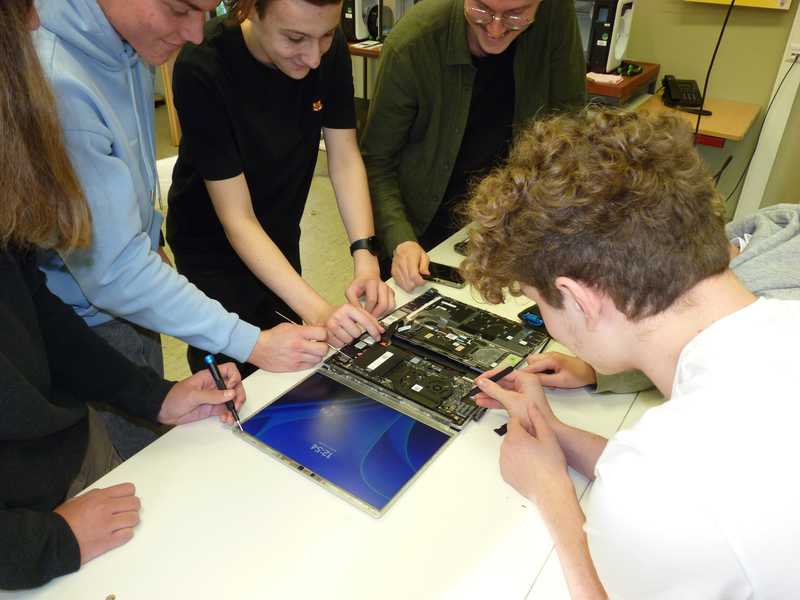 Eine Gruppe von Schülerinnen und Schülern beugt sich mit Werkzeugen in der Hand über das Innere eines Notebooks.