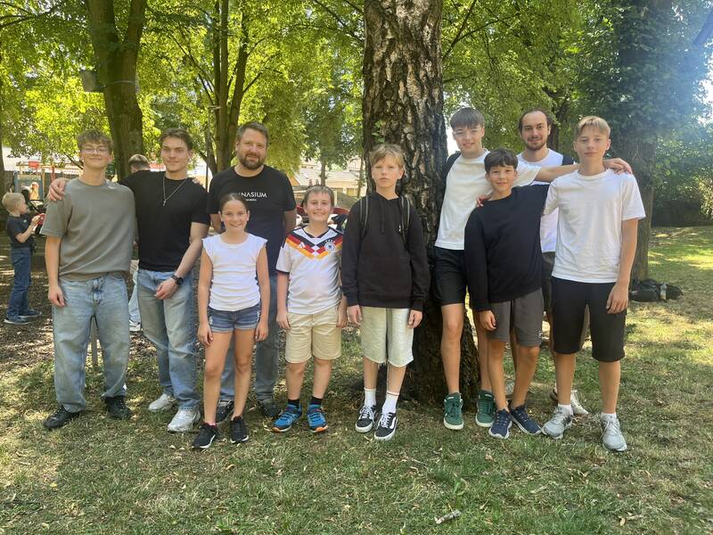 Eine Gruppe von Menschen, bestehend aus Erwachsenen und Kindern, steht fröhlich in einem parkähnlichen Gelände vor einem Baum.
