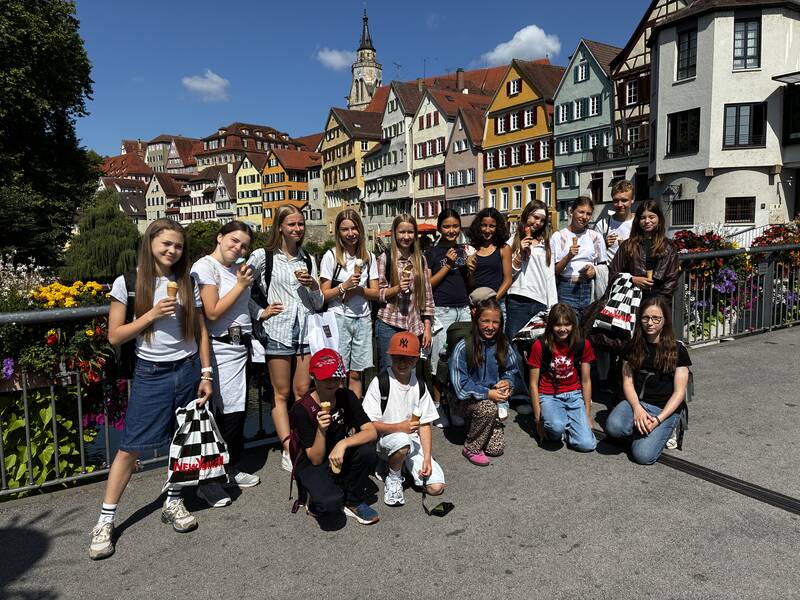 Eine Gruppe junger Menschen steht auf einer Brücke in einer malerischen Stadt mit bunten Fachwerkhäusern im Hintergrund und genießt Eiscreme.