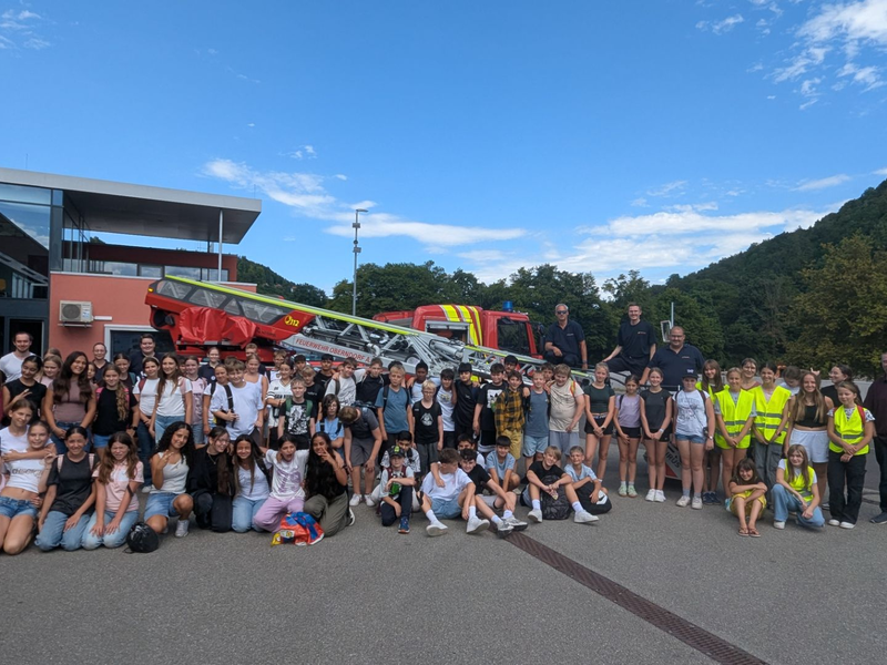 Eine große Gruppe von Menschen, darunter Kinder und Erwachsene, posiert gemeinsam vor einem Feuerwehrfahrzeug im Freien an einem sonnigen Tag.